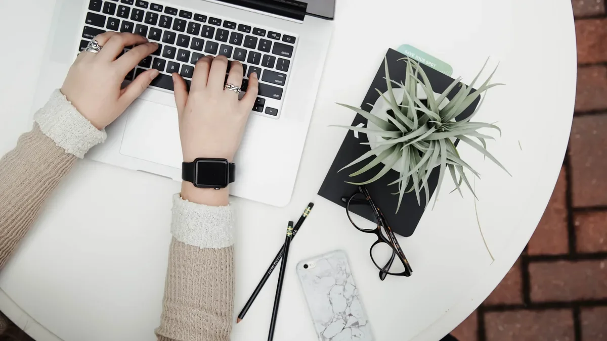 Typing on a laptop at a white table beside a notebook and an air plant