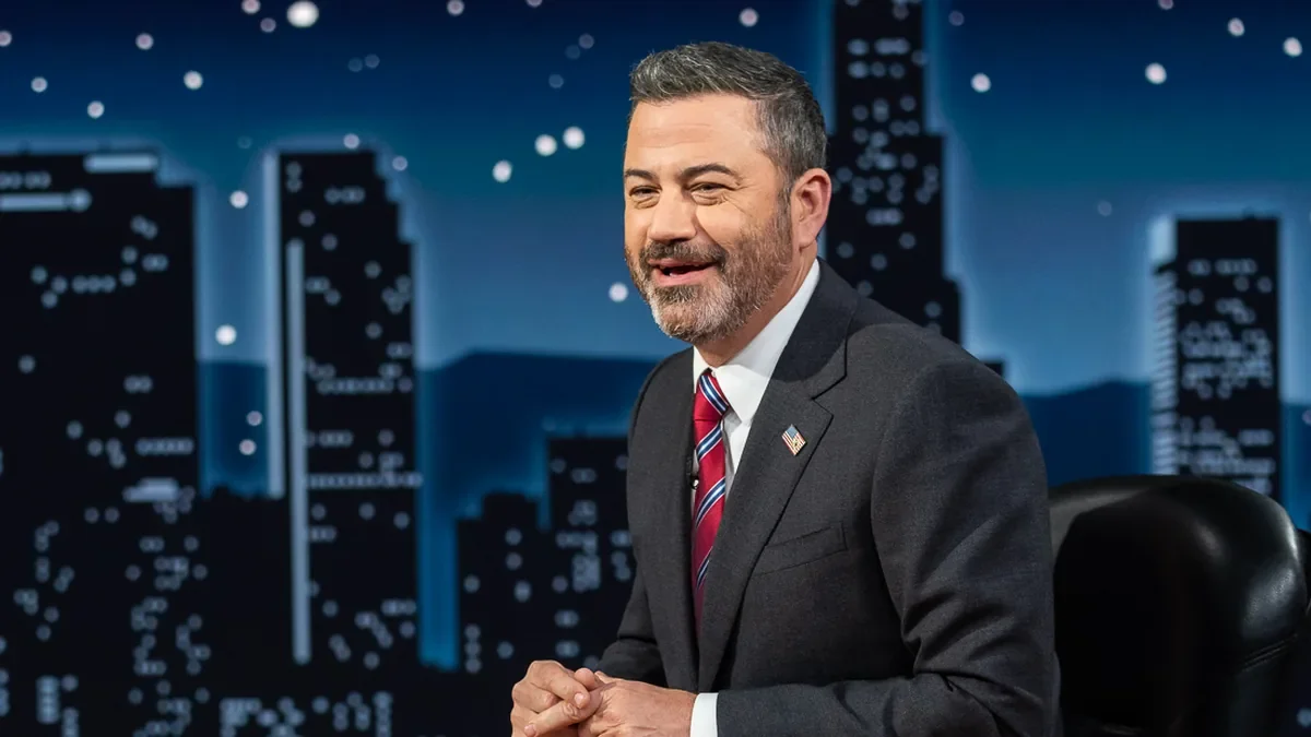 Late-night television host smiling on a talk-show set with a city skyline backdrop